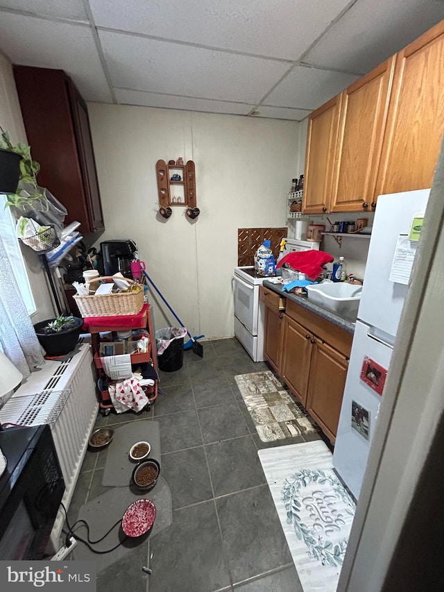kitchen featuring brown cabinets, a sink, dark tile patterned flooring, white appliances, and a paneled ceiling