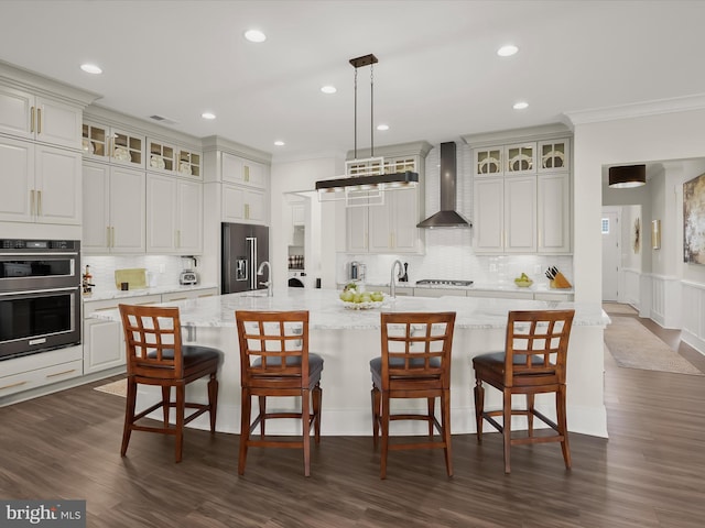 kitchen with crown molding, multiple ovens, wall chimney exhaust hood, a large island with sink, and high end refrigerator