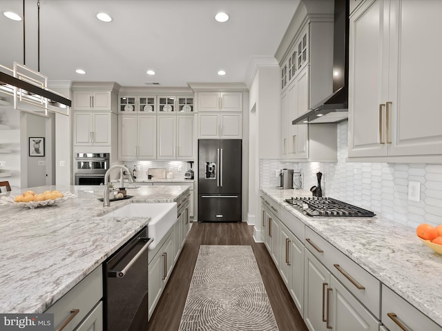 kitchen featuring wall chimney range hood, light stone countertops, a sink, stainless steel appliances, and dark wood-style floors