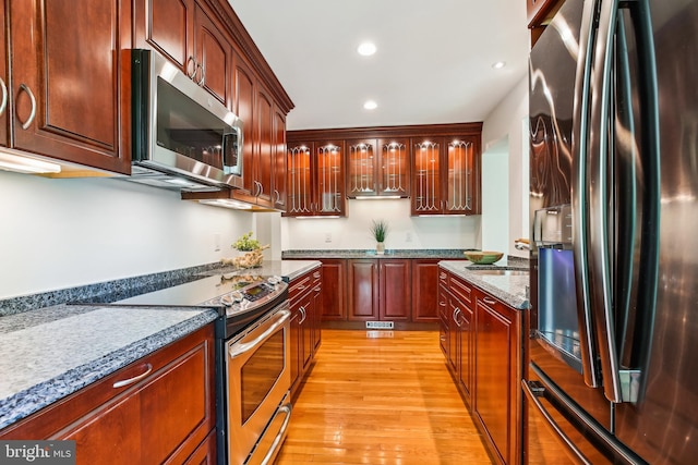 kitchen with glass insert cabinets, recessed lighting, light stone counters, light wood-style flooring, and stainless steel appliances