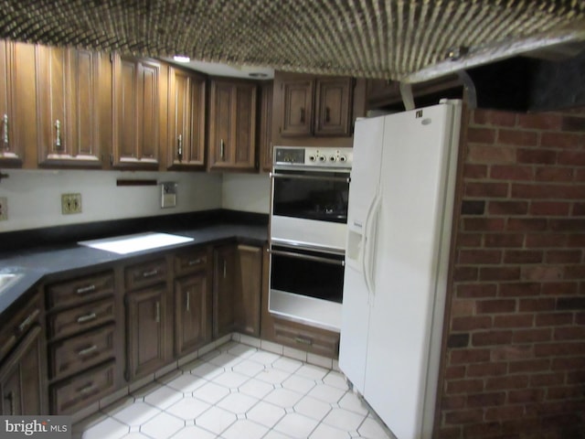 kitchen featuring dark countertops, dark brown cabinets, multiple ovens, light tile patterned floors, and white fridge with ice dispenser