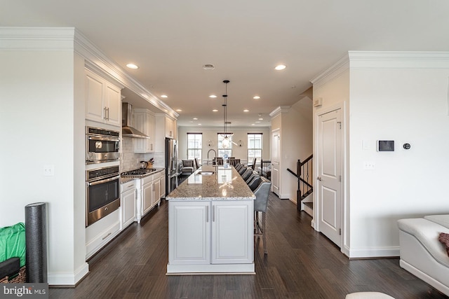 kitchen featuring wall chimney range hood, a sink, appliances with stainless steel finishes, recessed lighting, and an island with sink