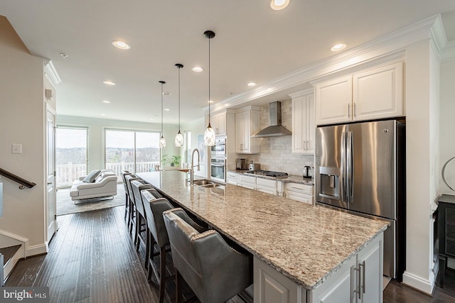 kitchen with a sink, a center island with sink, wall chimney exhaust hood, white cabinetry, and appliances with stainless steel finishes