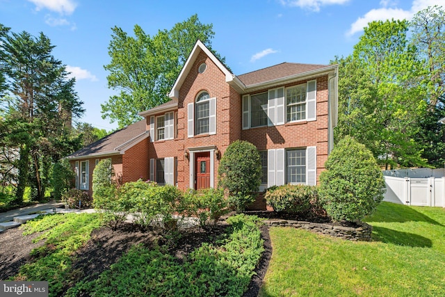 colonial-style house with fence, a shingled roof, a front lawn, brick siding, and a gate