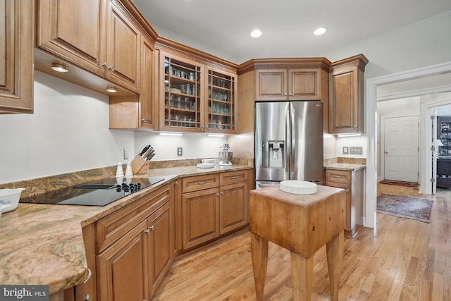 kitchen featuring black electric stovetop, brown cabinetry, light wood-style flooring, stainless steel refrigerator with ice dispenser, and glass insert cabinets