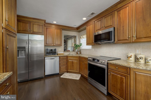 kitchen featuring appliances with stainless steel finishes, brown cabinetry, light stone counters, and dark wood-type flooring
