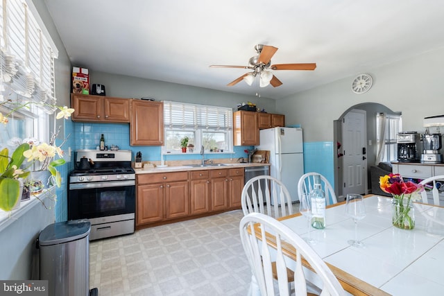 kitchen with a sink, stainless steel gas stove, brown cabinetry, freestanding refrigerator, and a ceiling fan