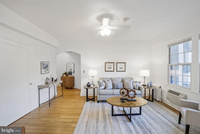 living room featuring radiator heating unit, ceiling fan, a wall unit AC, arched walkways, and light wood-style floors