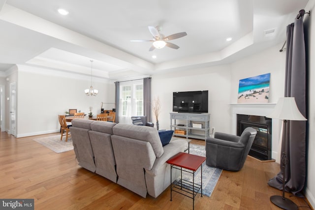 living area with light wood-style flooring, a raised ceiling, ceiling fan with notable chandelier, and a glass covered fireplace