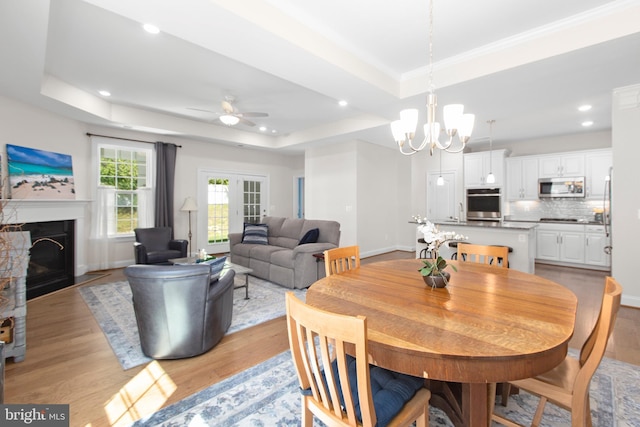 dining room featuring ceiling fan with notable chandelier, a tray ceiling, light wood finished floors, and a fireplace with flush hearth