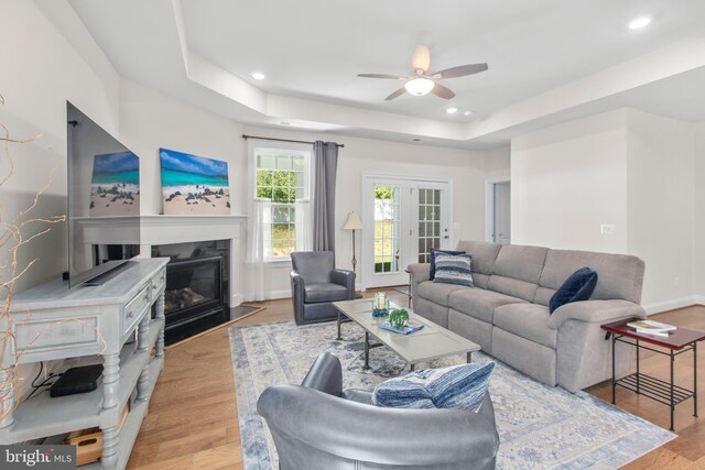 living area with light wood-style floors, ceiling fan, recessed lighting, a tray ceiling, and a fireplace with flush hearth