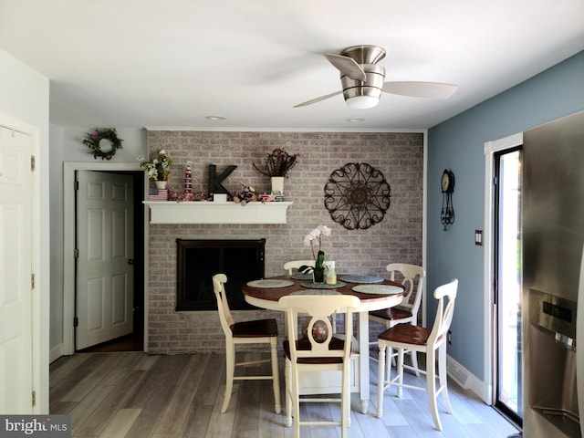 dining space featuring a fireplace, brick wall, ceiling fan, baseboards, and wood finished floors