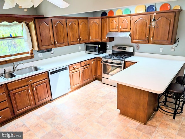kitchen featuring exhaust hood, brown cabinetry, appliances with stainless steel finishes, a sink, and a peninsula