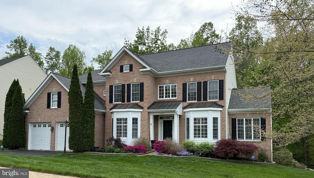 traditional-style home with aphalt driveway, roof with shingles, a garage, brick siding, and a front lawn