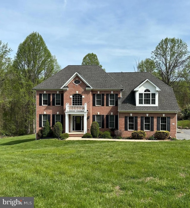 colonial home with a balcony, roof with shingles, a front lawn, and brick siding