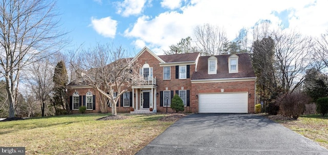 georgian-style home featuring brick siding, aphalt driveway, a balcony, a front yard, and an attached garage