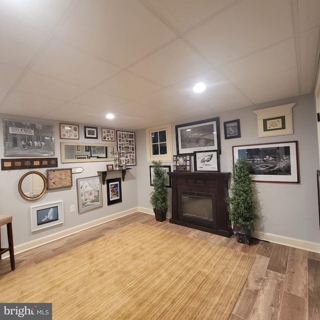living area with a glass covered fireplace, a drop ceiling, wood finished floors, and baseboards