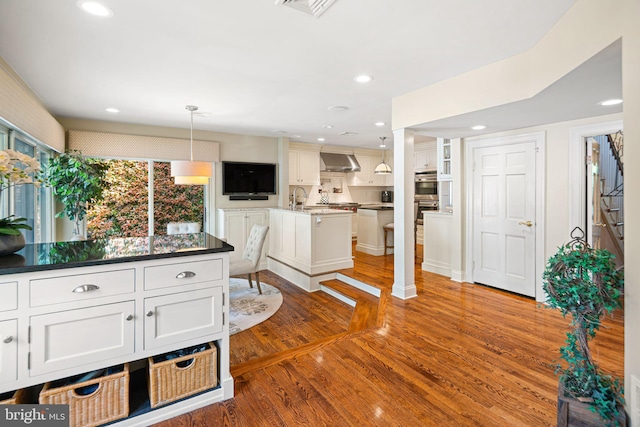 kitchen with wall chimney exhaust hood, wood finished floors, white cabinetry, pendant lighting, and stainless steel double oven