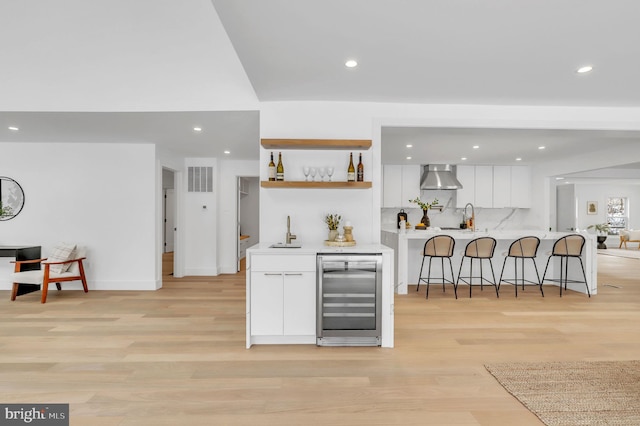 bar with wall chimney range hood, light wood finished floors, beverage cooler, recessed lighting, and wet bar