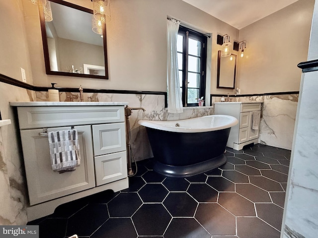 full bathroom with tile walls, wainscoting, a soaking tub, two vanities, and tile patterned flooring