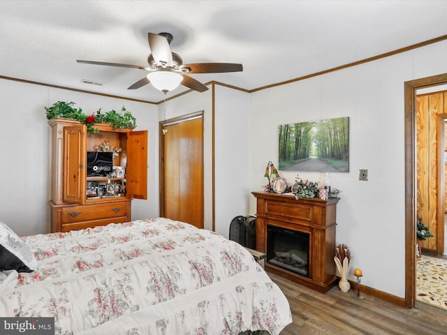 bedroom featuring ceiling fan, a glass covered fireplace, wood finished floors, visible vents, and ornamental molding