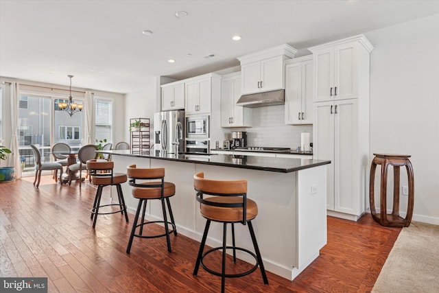 kitchen with a kitchen island with sink, dark wood-style flooring, stainless steel appliances, a chandelier, and under cabinet range hood