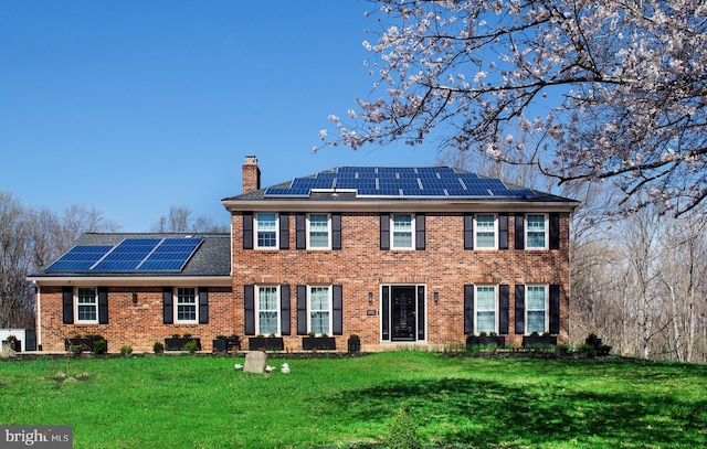 view of front of property featuring a chimney, brick siding, solar panels, roof with shingles, and a front yard