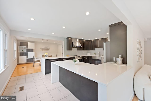 kitchen with wall chimney range hood, dark brown cabinets, a sink, a peninsula, and visible vents