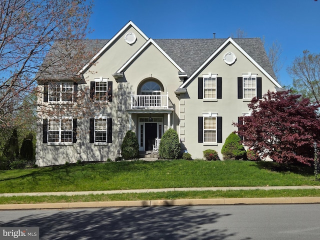 colonial house with stucco siding, a balcony, a shingled roof, and a front lawn