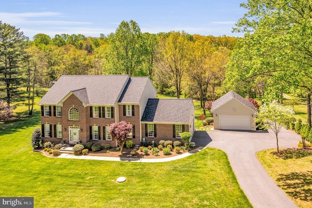 colonial house featuring a forest view, a detached garage, brick siding, a front lawn, and a shingled roof