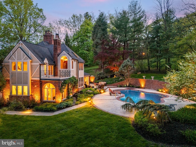 back of house at dusk featuring a balcony, a chimney, brick siding, a patio area, and a yard