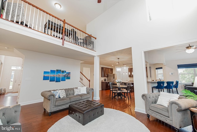 living area with a ceiling fan, stairway, baseboards, a towering ceiling, and dark wood-style floors