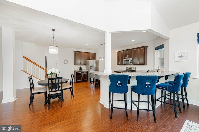 kitchen with dark wood finished floors, dark brown cabinetry, a kitchen bar, stainless steel appliances, and ornate columns