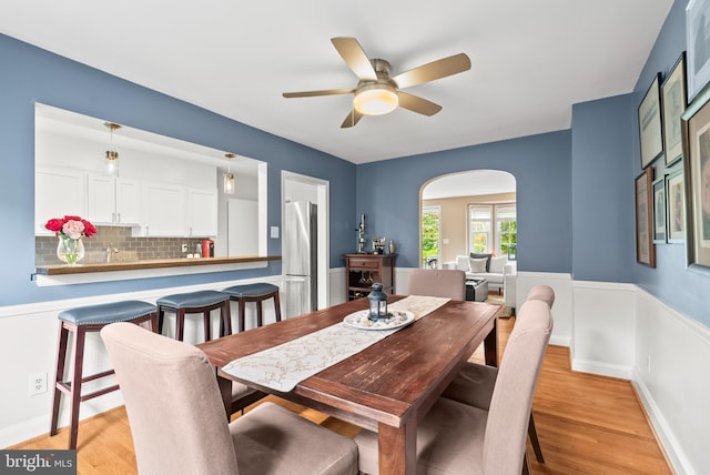dining room with arched walkways, ceiling fan, light wood-style flooring, and a wainscoted wall