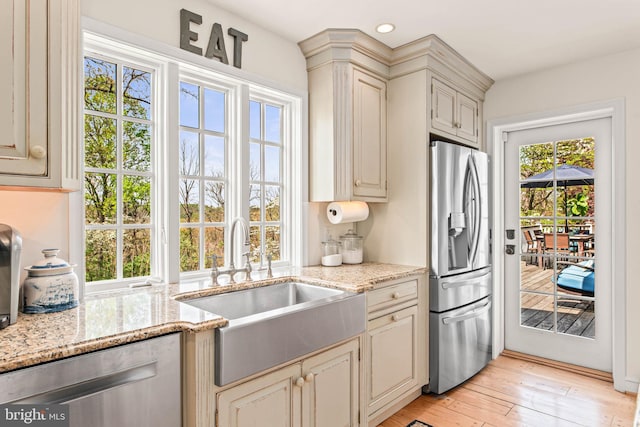 kitchen with appliances with stainless steel finishes, cream cabinets, a sink, light stone counters, and light wood finished floors