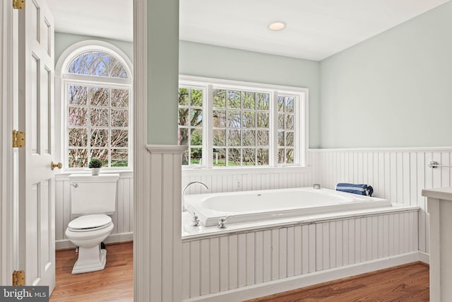 full bath featuring a garden tub, wood finished floors, a wainscoted wall, toilet, and a wealth of natural light
