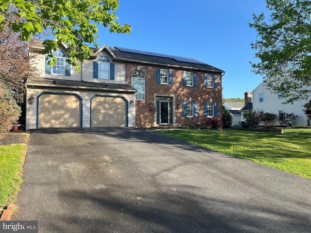 colonial house with an attached garage, brick siding, a front yard, solar panels, and driveway