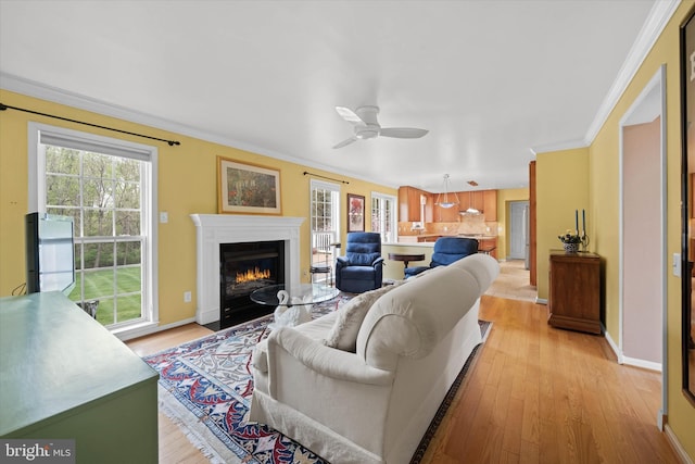 living area featuring a fireplace with flush hearth, crown molding, light wood-style flooring, baseboards, and a ceiling fan