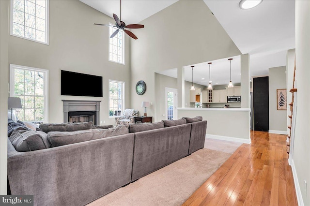 living area featuring light wood finished floors, a fireplace, a ceiling fan, baseboards, and a towering ceiling