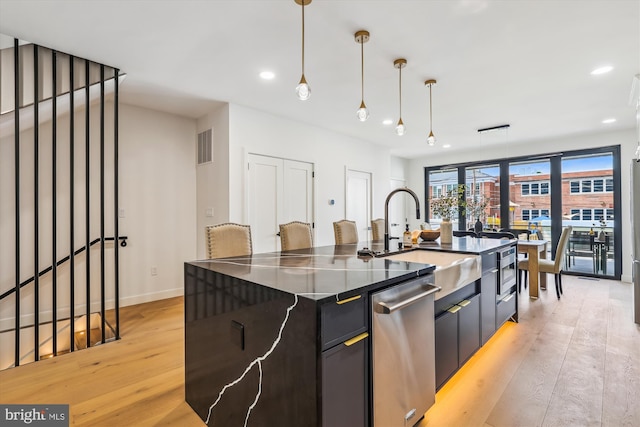 kitchen with stainless steel dishwasher, a sink, light wood-type flooring, a kitchen island with sink, and hanging light fixtures
