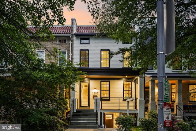 back of property at dusk featuring covered porch, stairway, a tile roof, and brick siding