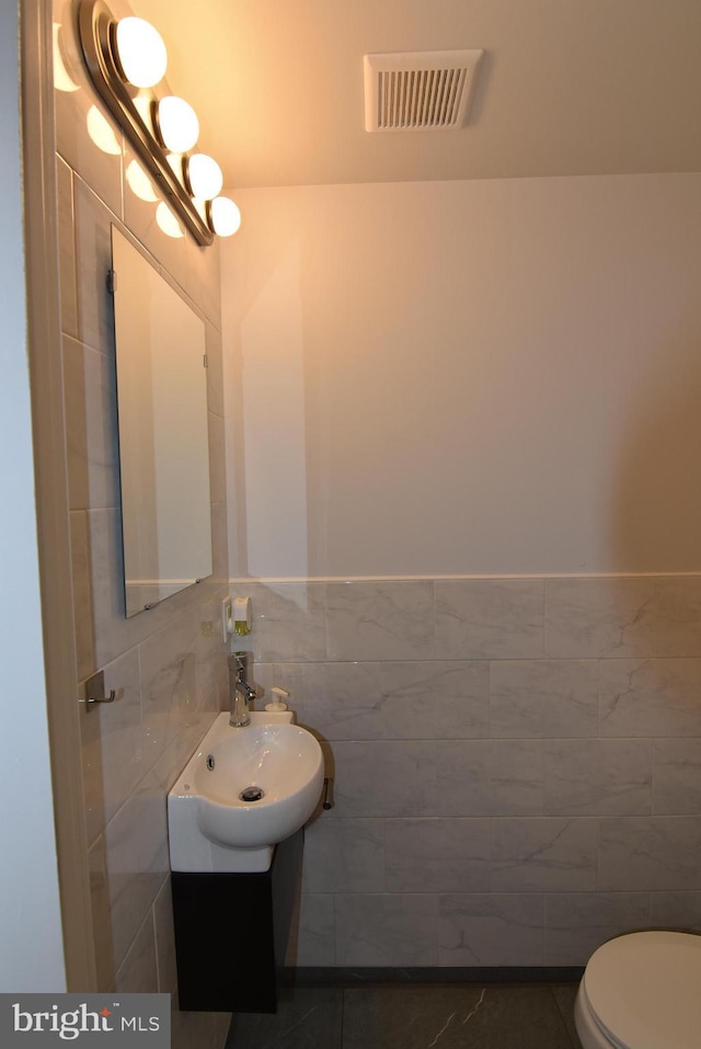 half bathroom featuring tile walls, a sink, toilet, visible vents, and wainscoting