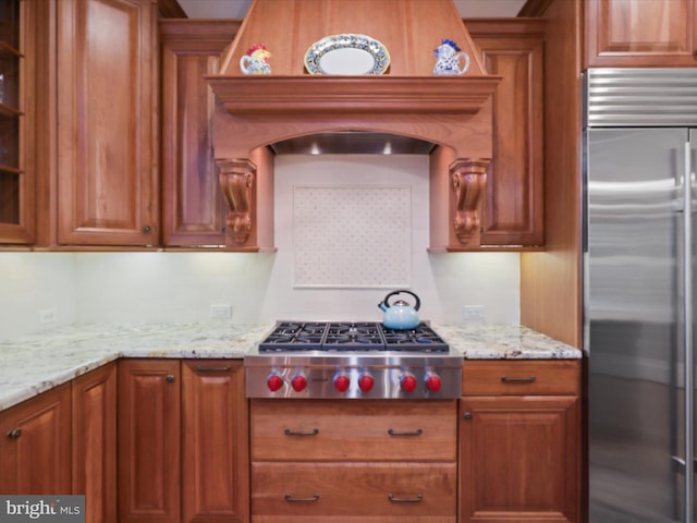 kitchen with glass insert cabinets, appliances with stainless steel finishes, brown cabinetry, and light stone counters