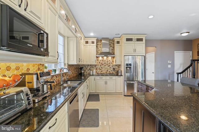 kitchen with wall chimney range hood, cream cabinets, a sink, appliances with stainless steel finishes, and backsplash