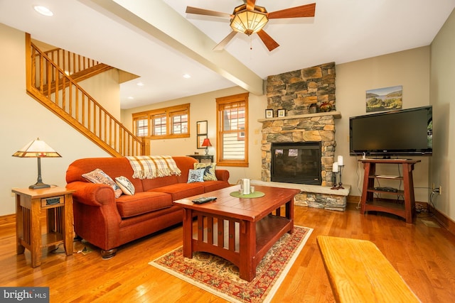 living room with beam ceiling, a stone fireplace, stairs, a ceiling fan, and wood finished floors