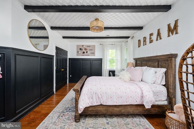 bedroom featuring dark wood-type flooring, wainscoting, a decorative wall, and beamed ceiling