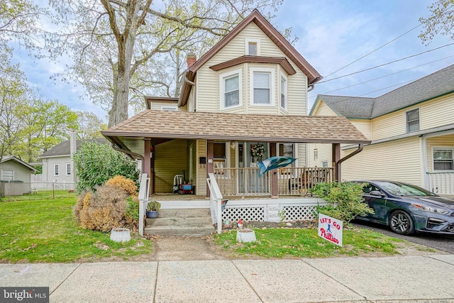 victorian home featuring covered porch, a chimney, a front yard, fence, and a shingled roof