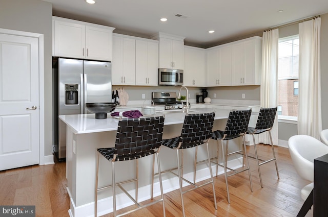 kitchen featuring light wood-style flooring, stainless steel appliances, a wealth of natural light, white cabinets, and a center island with sink