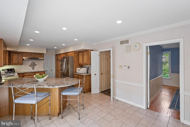 kitchen featuring brown cabinetry, crown molding, white microwave, a peninsula, and under cabinet range hood