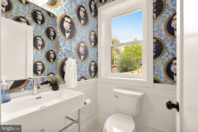 half bath featuring wainscoting, a sink, toilet, a decorative wall, and wallpapered walls